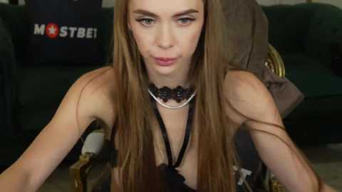 Media: Video of a young woman with long, straight brown hair and fair skin, wearing a black halter top, sitting on a couch with a plush brown pillow.