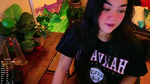 Media: Video of a young woman with long black hair, wearing a black \"AVAR\" t-shirt, sitting at a wooden table in a colorful, plant-filled room.