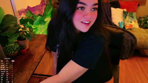 Media: A young woman with long black hair, fair skin, and dark eyes sits on a wooden chair in a cozy living room. She wears a black t-shirt and smiles at the camera.