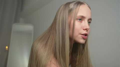 Media: Video of a young Caucasian woman with long, straight, blonde hair, fair skin, and blue eyes, wearing a neutral expression, standing in a softly lit room with a white lamp in the background.