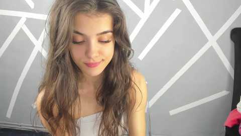 Media: Video of a young, fair-skinned woman with wavy brown hair and closed eyes, wearing a white strapless top, standing against a geometric, white-patterned gray wall.