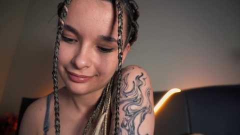 Media: Video of a young woman with light skin and intricate braided hair, smiling. She has a large tribal tattoo on her shoulder. The background is a dimly lit room with a couch and a warm, ambient light.