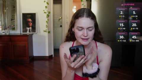 Media: A young woman with fair skin and long brown hair, wearing a red bra, takes a selfie with a smartphone, holding a stuffed animal in her right hand. Background shows a living room with a wooden cabinet, green ivy, and a framed photo of a man.