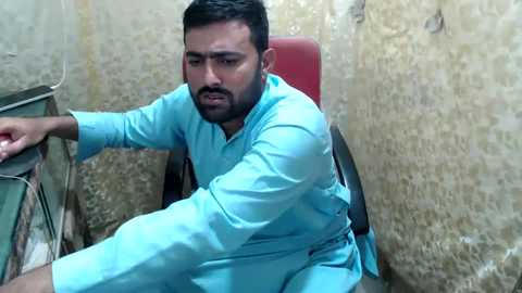 Media: Video of a man with short black hair and beard, wearing a light blue kurta, seated in a cramped, dimly lit room with a red chair and beige walls.