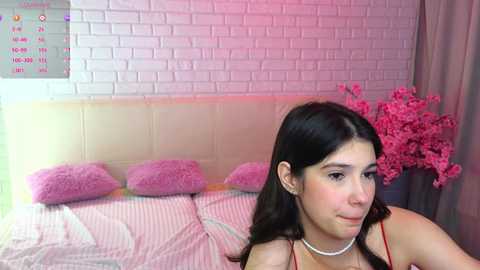 Media: Video of a young woman with long dark hair and fair skin, wearing a red dress and pearl necklace, sitting on a bed with pink pillows and a white brick wall background.