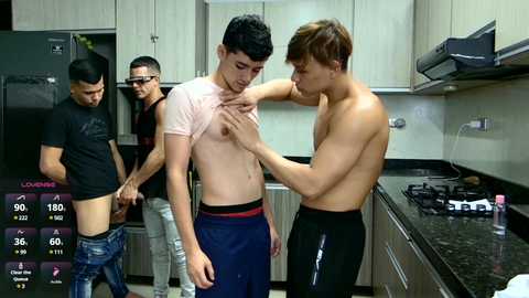 Media: Video: Two shirtless, young, fit men in a modern kitchen; one lifting the other's shirt, revealing his chest. Background: Wooden cabinets, black countertop, stove, and a third man in glasses and a black shirt.