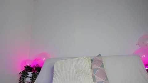 Media: Video of a white wall with pink and purple balloon lights, a white bed with a quilt featuring pastel geometric patterns, and a small green plant in the corner.