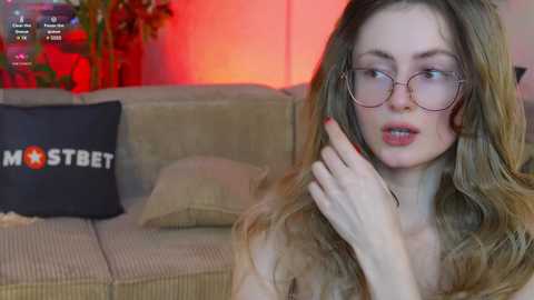 Media: Video of a pale-skinned woman with long, wavy blonde hair, wearing glasses, and a puzzled expression, sitting on a beige sofa with a black cushion featuring \"MOTHER\" text.