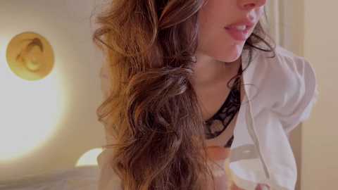 Media: Video of a young woman with long, wavy brown hair styled in pigtails, wearing a white hoodie and black lace bra, partially seen. Background shows a softly lit, beige wall with a circular light fixture.