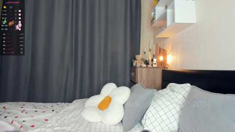 Media: Video of a modern bedroom with a large bed, white floral pillow, grey curtains, and wooden nightstand with a lamp, calendar, and decorative items.