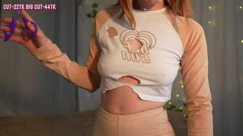 Media: Video of a young woman with light skin and long brown hair wearing a white and peach crop top with \"HOT\" written on it, holding purple scissors. Background has blurred curtains and green plants.