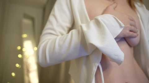 Media: A video captures a topless woman with light skin, wearing a white, long-sleeved cardigan, exposing her medium-sized breasts. The background is blurred, featuring a soft, ambient light.