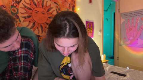 Media: Video of a young woman with long hair leaning over a man with short hair, both wearing casual clothes, in a colorful, eclectic bedroom with sunburst tapestry, posters, and colorful lights.