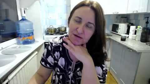 Media: Video of a woman with shoulder-length brown hair, wearing a black floral shirt, sitting in a modern kitchen with white cabinets, white countertop, and a blue water jug.