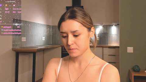 Media: Video of a young woman with fair skin and brown hair in a ponytail, wearing a white tank top, sitting in a modern, beige kitchen with tiled backsplash, and a light bulb hanging.