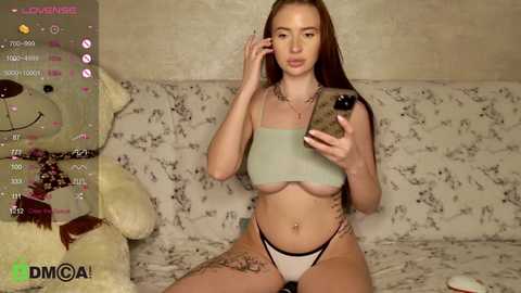 Media: Video of a young Caucasian woman with long brown hair, wearing a light blue crop top and white thong, sitting on a floral-patterned couch, holding a phone, in a cozy room with a teddy bear.