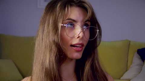 Video of a young woman with long, straight brown hair, wearing round glasses, and sitting on a mustard-yellow couch in a dimly lit room.