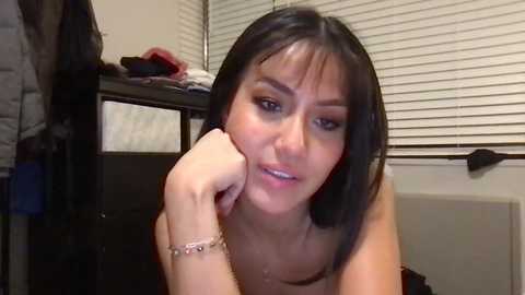 Media: Video of a young Asian woman with straight black hair, light skin, and dark eyes, wearing minimal makeup. She's leaning on her elbow, looking introspective. Background includes a dark dresser with clothes and white blinds.