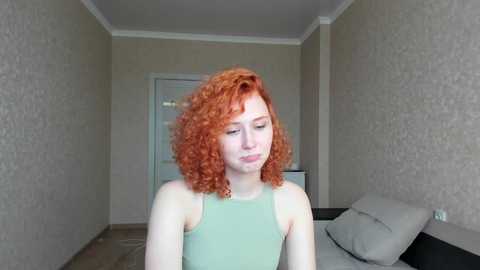 Media: Video of a fair-skinned, young woman with curly, vibrant red hair, wearing a sleeveless mint green top, standing in a beige-tiled room with patterned wallpaper and a white door in the background.