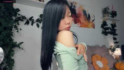 Media: Video of an Asian woman with long black hair, wearing a light green off-shoulder sweater, standing in a cozy, decorated room with floral artwork and greenery.