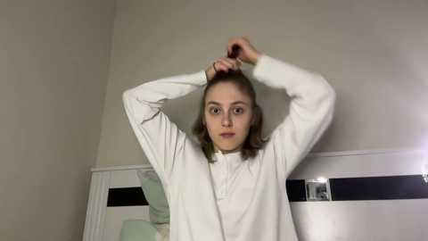 Media: Video of a young Caucasian girl with shoulder-length brown hair, wearing a white long-sleeve shirt, adjusting her hair in a bedroom with white walls and a mirrored dresser.