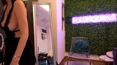 Media: Video of a woman with fair skin and long dark hair in a black, sleeveless dress, standing next to a full-length mirror in a room with a lush green wall and a neon \"Silhouette\" sign.