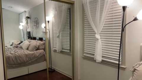 Media: Video of a minimalist, modern bedroom with a white bed adorned with beige pillows, a white window blind, and a tall floor lamp.