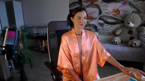 Media: Video of a woman in a light pink robe sitting in a black chair, surrounded by a cozy bedroom with a plush teddy bear, a plant, and a bookshelf in the background.