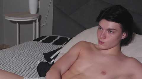 Media: Video of a shirtless, fair-skinned, young man with short dark hair, lying on a bed with a gray and white patterned blanket. Background shows a white bedside table and a gray headboard.