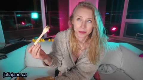 Media: Video of a blonde woman in a light-colored robe, holding a fork, with a surprised expression, in a modern, dimly-lit room with neon lighting and a balcony view.