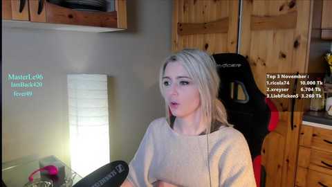 Media: Video of a blonde woman in a beige sweater, seated at a wooden desk with a lamp, holding a microphone, in a wooden-paneled room, with text overlay.