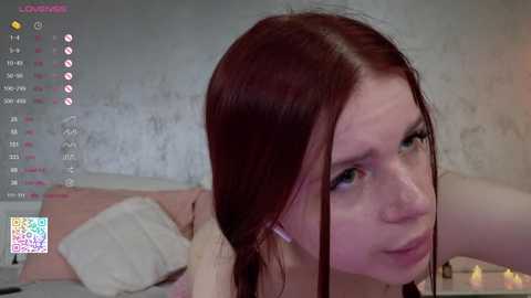 Media: Video of a pale-skinned woman with red hair, looking pensive, lying on a bed with white pillows. Background shows a dimly lit room with a textured wall and a candle on a nightstand.