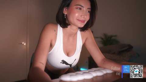 Media: A video of a fit woman with short black hair, wearing a white sports bra and shorts, working out with a foam roller. Background shows a blurred room with a bed and a plant.