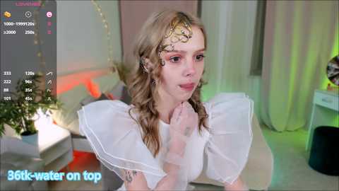 Media: Video of a young woman with blonde hair, wearing a white sheer top with ruffled sleeves, standing in a brightly lit, modern room with green and red accents, looking at her phone.