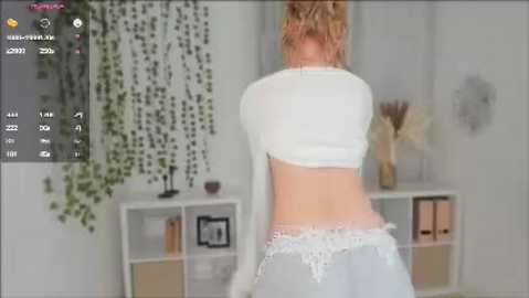 Media: Video of a woman in a white crop top and lace-trimmed jeans, standing with her back to the camera, in a modern, minimalist bedroom with green vines, white shelves, and decor.