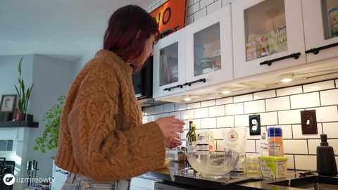 Media: Video of a woman with red hair, wearing a brown cable-knit sweater, cooking in a modern kitchen with white cabinets, black handles, and a white subway tile backsplash.