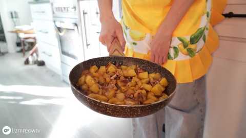 Media: Video of a person in a yellow apron, holding a skillet with yellow potatoes and bacon, in a modern kitchen with white cabinets and a grey countertop.