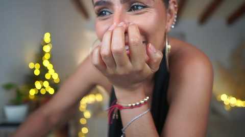 Media: Video of a smiling woman in a black dress, covering her mouth with her hands, wearing gold hoop earrings and bracelets. Background features a blurred Christmas tree with twinkling lights.