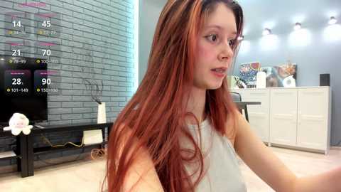 Media: Video of a young woman with long, red hair in a white tank top, sitting in a modern living room with a brick wall, flat-screen TV, and white cabinets.