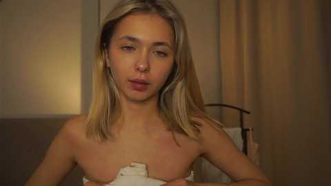 Media: Video of a topless young woman with light skin, straight blonde hair, and minimal makeup, sitting in a dimly lit room with beige curtains and a bed.