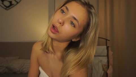 Media: Video of a young, light-skinned woman with long, blonde hair, wearing a white towel, leaning forward, eyes closed, in a dimly lit bedroom.