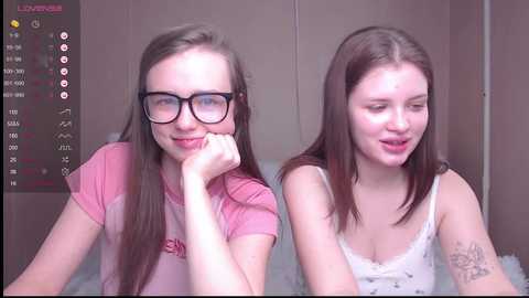 Media: Video of two young Caucasian women with fair skin, one with glasses, in casual clothes, smiling at each other, set in a bathroom with a digital display.