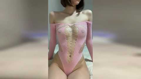 Media: Video of a slender, fair-skinned woman with short brown hair, wearing a revealing, pink, lace-up bodysuit. The background is a blurred, beige-toned room with soft lighting.