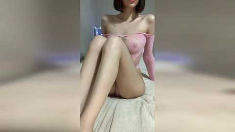 Media: Video of a slender Asian woman with short brown hair, wearing a pink off-shoulder top and revealing her small breasts, sitting on a bed with beige sheets. Background is a dimly lit, plain room with a blue object on the left.