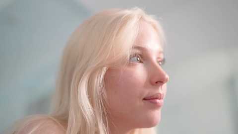 Video of a young Caucasian woman with light blonde hair, fair skin, and blue eyes, gazing thoughtfully to the right. She wears a soft, natural makeup look, and the background is a blurred, pale blue and white gradient.