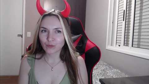 Media: Video of a young woman with light skin and long blonde hair, wearing a green tank top and red devil horns. She sits in a black and red gaming chair with white blinds in the background.