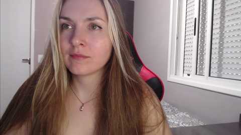 Media: Video of a young woman with long, straight, blonde hair, fair skin, and light makeup. She's wearing a necklace and is seated indoors near a window with white blinds.