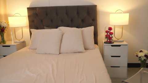 Media: Video of a modern, minimalist bedroom with a plush, gray tufted headboard, white linens, two white nightstands, and matching white drawers. The room is warmly lit by two white table lamps, with a vase of red roses on one nightstand.