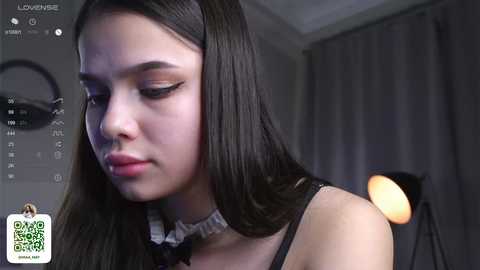 Media: Video of a young Asian woman with long black hair and light skin, wearing a black lace top with a white bow, looking contemplative in a dimly lit room with a lamp in the background.