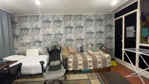 Media: Video of a small, sparsely furnished room with a gray and white patterned wallpaper, featuring a black gaming chair, a bed with a plaid blanket, and a white desk with a chair.